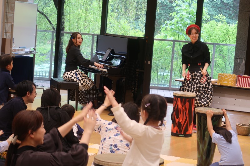 大きな窓から緑が見える部屋で、ピアノと打楽器を演奏する2人の女性と、手振りをして楽しむ参加者の様子。