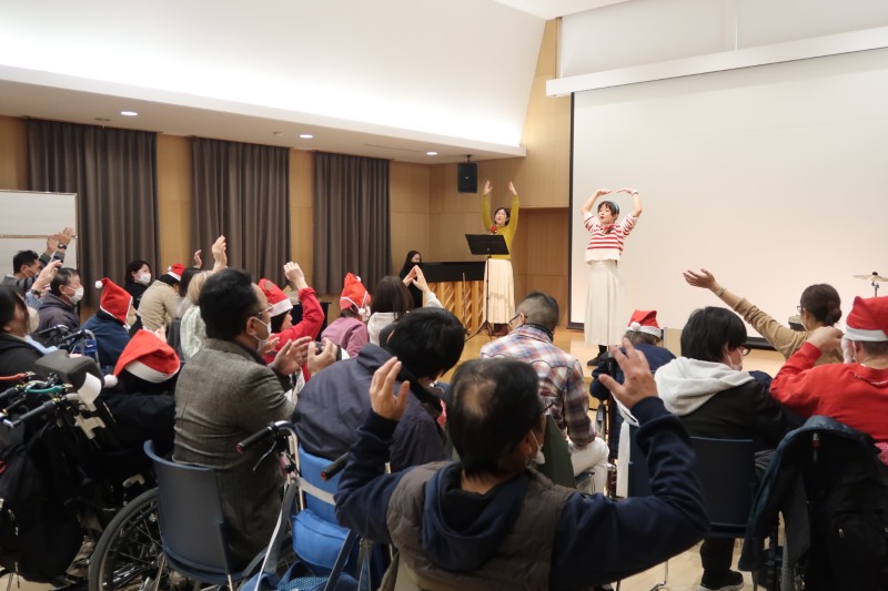 サンタ帽をかぶった観客席の人々が、ステージ上の2人の女性パフォーマーに合わせて手を挙げている、イベントの様子。
