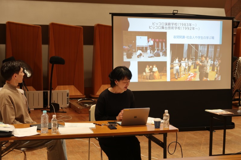 ピッコロ演劇学校に関するスライドを背に、ノートパソコンを前にして話をする女性講師と、それを横で見守る男性の様子。