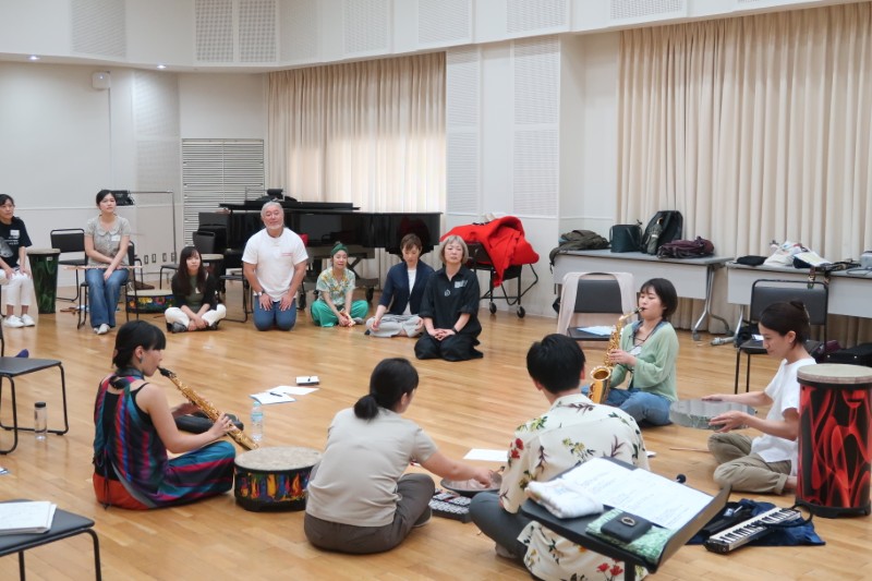 明るい練習室で、サックスや太鼓を演奏する人と、床に座ってそれを見つめる参加者たちが大きな円を作っている様子。